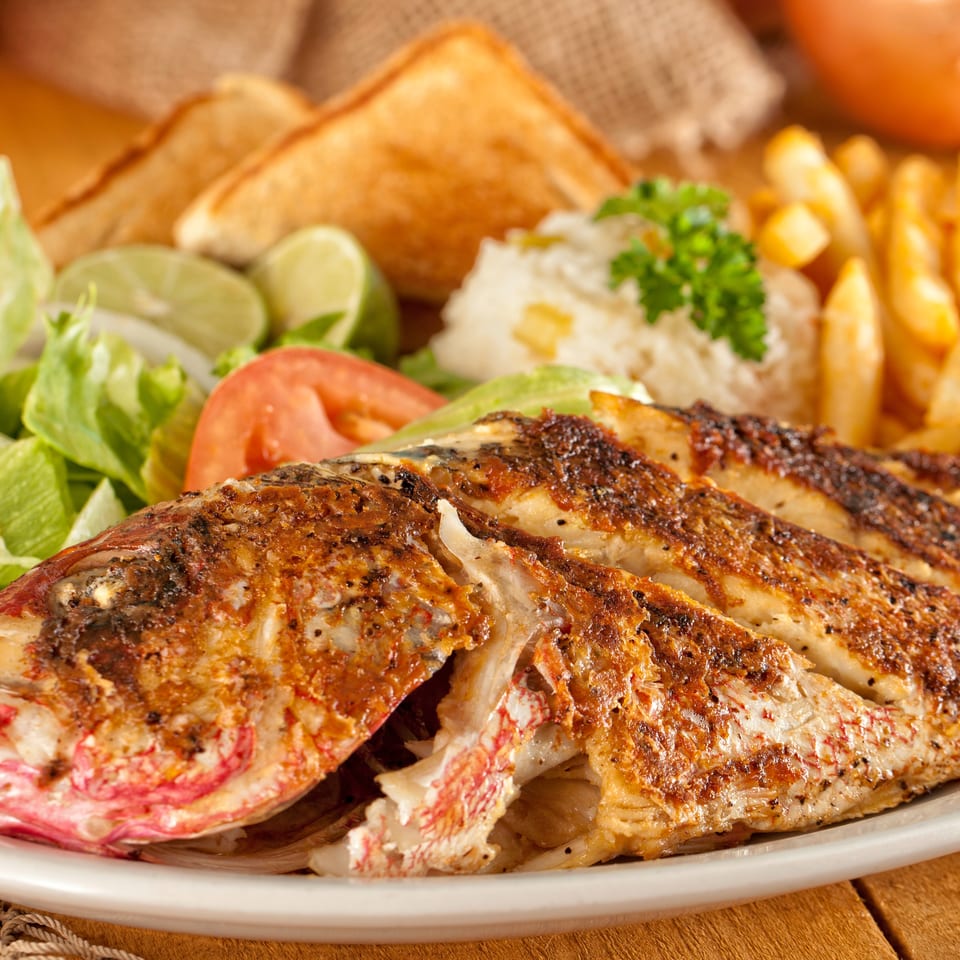 Grilled whole fish served with fries, rice, salad, lime wedges, and toasted bread on a restaurant platter.
