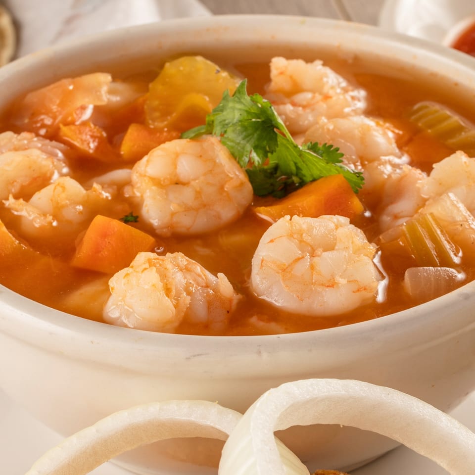 Bowl of shrimp soup with vegetables and cilantro, served with tortillas, salsa, and white onion rings.