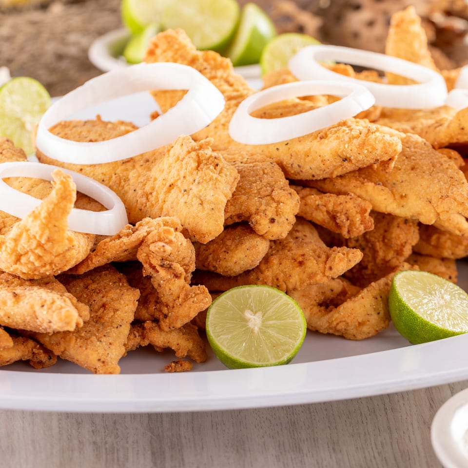 Golden fried fish piled on a platter with white onion rings, lime wedges, and dipping sauce.