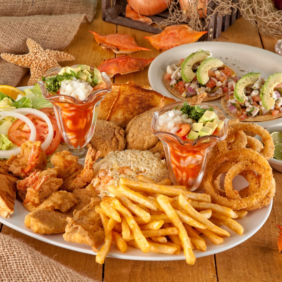Seafood platter with fried shrimp, fish, fries, rice, onion rings, ceviche, avocado, salad, and cocktail sauce on a rustic table.