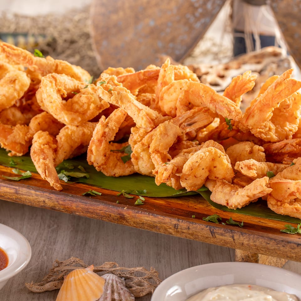 Crispy fried shrimp piled on a wooden platter lined with a banana leaf, accompanied by dipping sauces and seashell decorations.