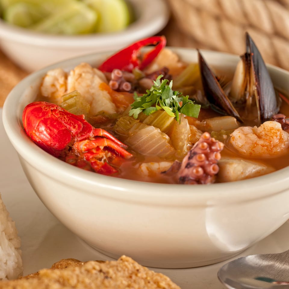Bowl of seafood soup with lobster, shrimp, mussels, octopus, vegetables, and herbs, served alongside rice.